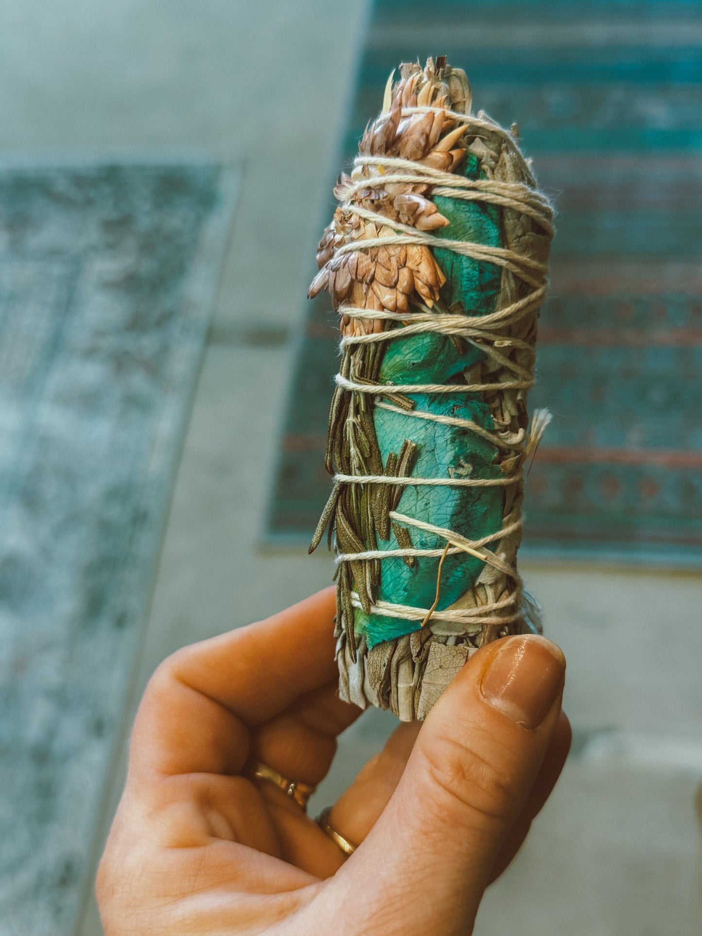 A hand holding a Flower Faerie Sage Smudge wand, with visible sage leaves and decorative flowers.